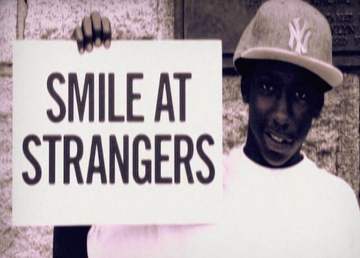 Man holding a sign that says "Smile at Strangers" with a New York Yankees cap in outdoor setting.