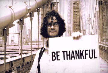 Man holding a sign with "BE THANKFUL'" text on bridge in outdoor setting. 