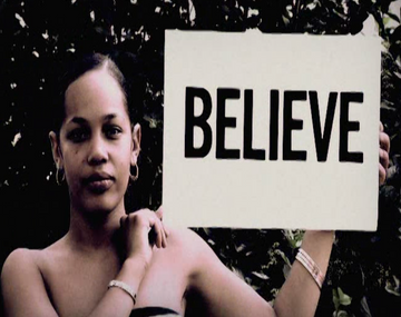 Woman holding a sign with the word "BELIEVE" against a dark background in outdoor setting. 