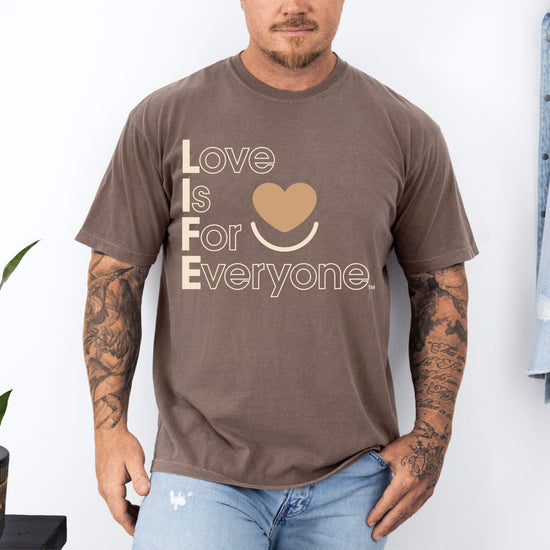 Man wearing a Comfort Colors espresso t-shirt with 'Love is for Everyone' text, standing against a white wall. 