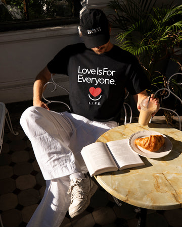 Man wearing Classic Tee in black, sitting at table showing the signature "Love is for Everyone" design with red heart and smile. 