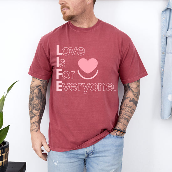 Man wearing Comfort Colors washed denim t-shirt with 'Love is for Everyone' text and heart symbol, standing against a white wall 