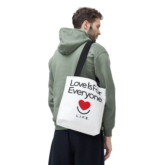 Man carrying white L.I.F.E. Signature Canvas tote with "Love is for Everyone" print and black handles. 