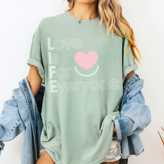 Woman wearing Bay pigment-dyed Comfort Colors ®  tee with "Love is for Everyone" heart graphic. 