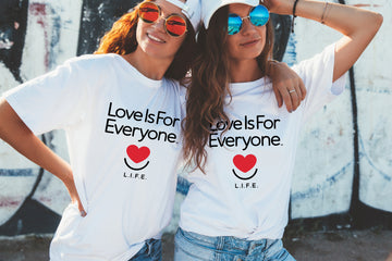 Two people wearing white t-shirts with "Love is for Everyone" text and heart smile logo, outdoor setting. 