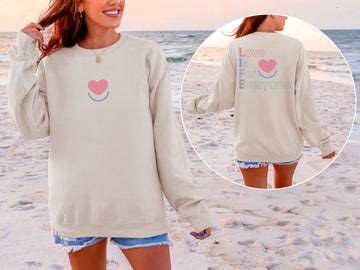 Woman wearing women's Color Pop Crewneck sand color, showing back "Love is for Everyone" message and front smiling heart design.